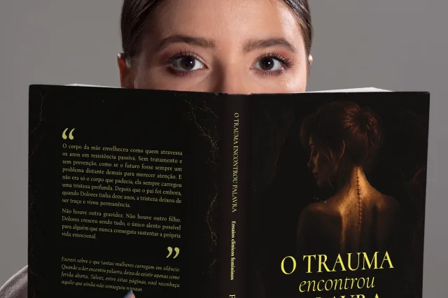 Leitora com o exemplar impresso de O Trauma encontrou Palavra aberto, mostrando a capa e a contracapa.